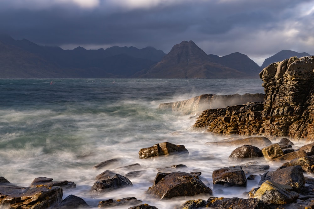 a rocky beach with a body of water and mountains in the background