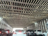 A traffic situation with multiple cars aligned in two lanes underneath a metal bridge with a grid-like pattern above. The bridge structure features criss-cross beams, and daylight is visible at the end of the tunnel-like passage. The vehicles are primarily white and black, with their tail lights illuminated.