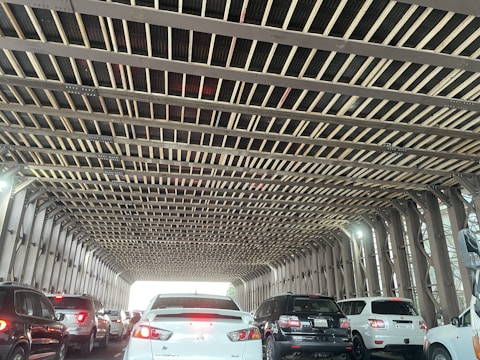 A traffic situation with multiple cars aligned in two lanes underneath a metal bridge with a grid-like pattern above. The bridge structure features criss-cross beams, and daylight is visible at the end of the tunnel-like passage. The vehicles are primarily white and black, with their tail lights illuminated.
