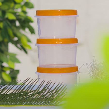 Three transparent plastic containers with orange lids are stacked vertically in an outdoor setting. Surrounding them are leaves and greenery, creating a fresh and natural ambiance.