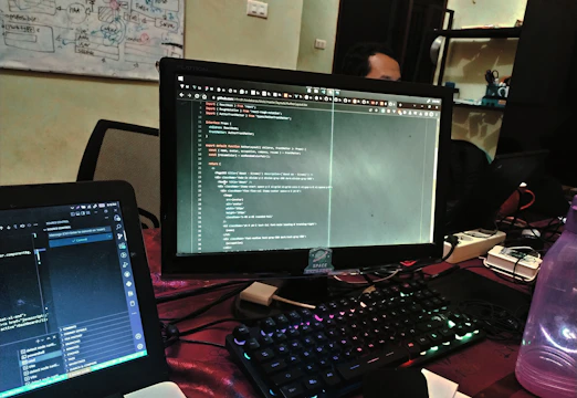 Close-up of hands typing code on a laptop with multiple monitors in the background.