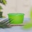 A bright green plastic container with a lid is placed on a wooden surface, surrounded by green foliage and a blurry background. The setting appears to be outdoors with natural lighting.