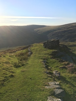 A serene landscape of green hills with a winding path leading up to a stone structure. The sky is clear with a soft gradient from blue to light yellow near the horizon. Sunlight gently illuminates the scene, casting long shadows.