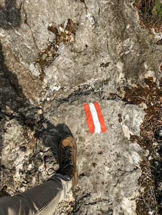 A hiking boot rests on a rocky terrain featuring a trail marker painted in red and white. The surface is rough with visible small cracks and patches of moss or dirt, indicating a natural outdoor setting.