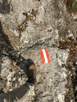 A hiking boot rests on a rocky terrain featuring a trail marker painted in red and white. The surface is rough with visible small cracks and patches of moss or dirt, indicating a natural outdoor setting.