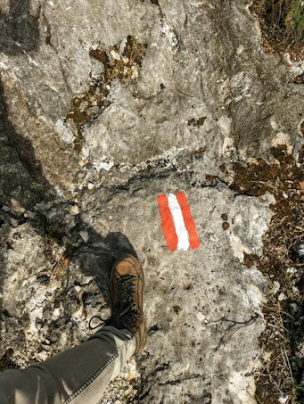 A hiking boot rests on a rocky terrain featuring a trail marker painted in red and white. The surface is rough with visible small cracks and patches of moss or dirt, indicating a natural outdoor setting.