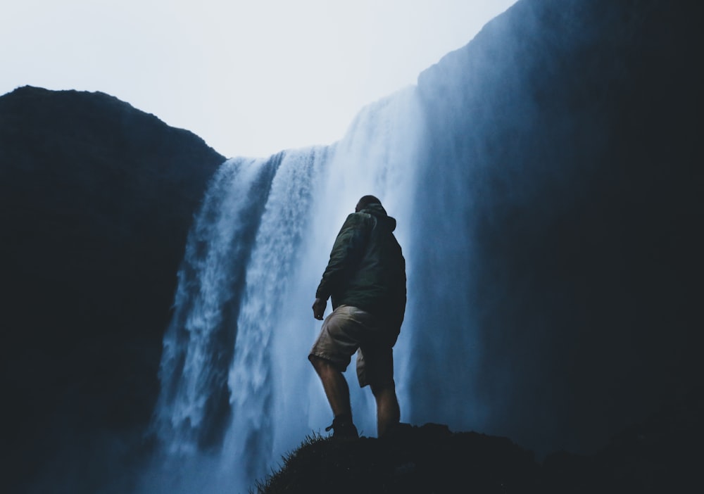 um homem de pé em uma rocha com uma cachoeira atrás dele