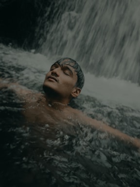 A person is relaxing in water with closed eyes, surrounded by the flowing movement of a waterfall. Water cascades down in the background and ripples form around the person in a serene, natural environment.