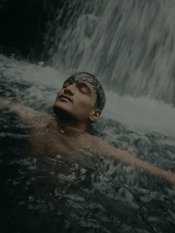A person is relaxing in water with closed eyes, surrounded by the flowing movement of a waterfall. Water cascades down in the background and ripples form around the person in a serene, natural environment.