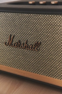 A close-up view of a vintage-style speaker with a woven fabric front and the brand name 'Marshall' prominently displayed in bronze lettering. The top of the speaker shows several metallic control knobs with a brushed finish.