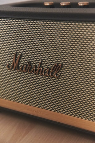 A close-up view of a vintage-style speaker with a woven fabric front and the brand name 'Marshall' prominently displayed in bronze lettering. The top of the speaker shows several metallic control knobs with a brushed finish.