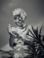A large geometric sculpture of a person is depicted in a monochromatic tone. The figure has a faceted, angular surface and holds a similarly styled object. The towering structure is seen against a backdrop of a building with angular architecture, and foliage partially obscures the lower right corner.