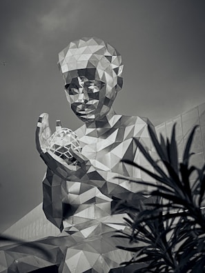 A large geometric sculpture of a person is depicted in a monochromatic tone. The figure has a faceted, angular surface and holds a similarly styled object. The towering structure is seen against a backdrop of a building with angular architecture, and foliage partially obscures the lower right corner.