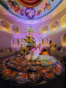 A vibrant setting with an ornate statue of a deity at the center, surrounded by colorful artwork. The ceiling is decorated with detailed paintings. Two people are present; one seated in front of the artwork, and another standing nearby.