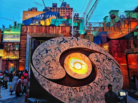 A large, circular mural with intricate black and white designs depicting various figures, possibly mythological, is the central focus. It appears to be an art installation with a glowing center, set against a vibrant backdrop of buildings adorned with colorful panels and vintage television antennas. There are several people gathered around, adding a sense of community or event.