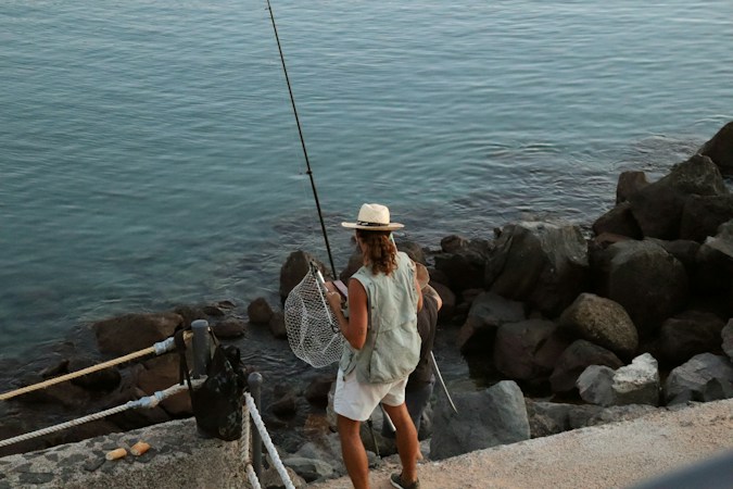 Angler in a straw hat and vest standing on a rock jetty above calm sea, holding a rod in one hand and a landing net in the other