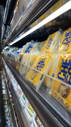 A supermarket shelf displays neatly organized packs of cheese in transparent plastic packaging. The yellow and blue design on the packaging stands out. Below these, a lower shelf holds containers of various dairy products. The lighting is bright, highlighting the products.
