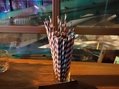 A collection of red and white striped paper straws is arranged in a clear glass on a wooden surface, with a glass of liquid on the left side. The background includes blurred reflections and a window, giving off colorful ambient lighting.