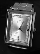 A rectangular silver wristwatch with a metallic bracelet and a minimalistic design. The watch face features a simple, silver-toned dial with black markers and hands. The brand name is displayed prominently at the center of the dial, and there is a date window at the six o'clock position.