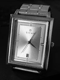 A rectangular silver wristwatch with a metallic bracelet and a minimalistic design. The watch face features a simple, silver-toned dial with black markers and hands. The brand name is displayed prominently at the center of the dial, and there is a date window at the six o'clock position.