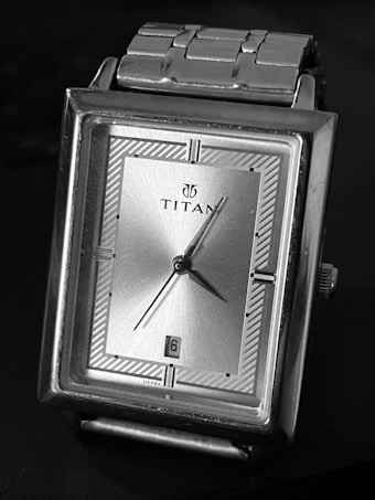 A rectangular silver wristwatch with a metallic bracelet and a minimalistic design. The watch face features a simple, silver-toned dial with black markers and hands. The brand name is displayed prominently at the center of the dial, and there is a date window at the six o'clock position.