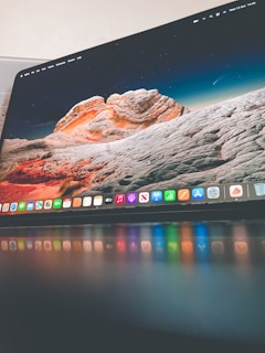 A sleek computer monitor displaying a colorful desktop background with a landscape of rocky formations under a starry sky. The foreground shows a glossy surface reflecting the vibrant colors of the desktop icons.
