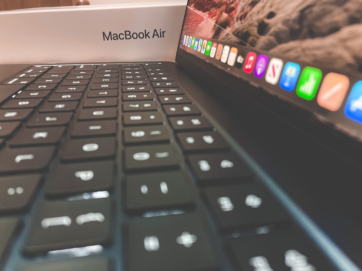 A close-up view of a laptop keyboard with a focus on the keys and the colorful application icons on the screen. The laptop is placed inside its packaging, which features the label 'MacBook Air'.