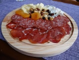 A wooden platter featuring an assortment of sliced cured meats, cubes of cheese, dried apricots, and a mix of nuts and berries. The arrangement is placed on a purple cloth background.