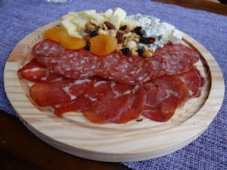 A wooden platter featuring an assortment of sliced cured meats, cubes of cheese, dried apricots, and a mix of nuts and berries. The arrangement is placed on a purple cloth background.