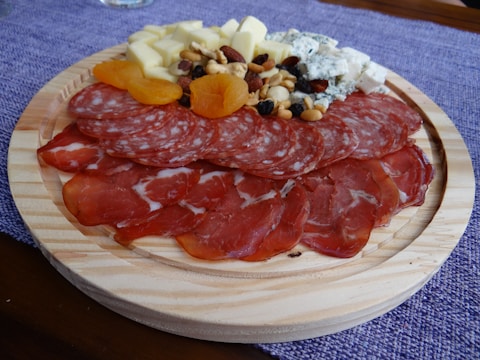 A wooden platter featuring an assortment of sliced cured meats, cubes of cheese, dried apricots, and a mix of nuts and berries. The arrangement is placed on a purple cloth background.