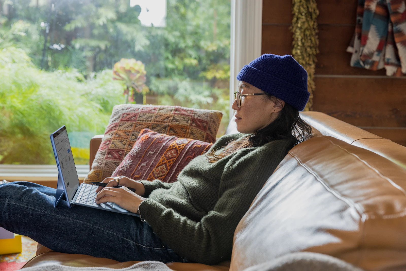 A woman in glasses and a knit beanie working on a laptop on a sofa by a garden window