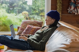 A person wearing a blue knit cap and glasses, sitting comfortably on a tan leather couch with a laptop in their lap. Several colorful, patterned cushions provide additional comfort. The background features a large window with a view of lush greenery, suggesting a relaxing and cozy atmosphere.