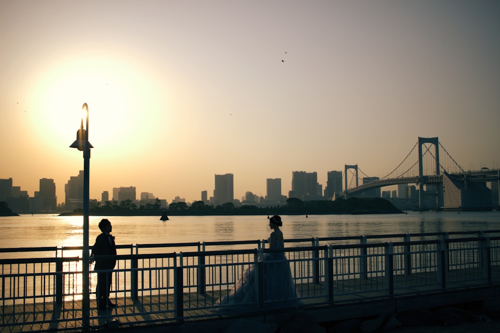 a couple of people standing on a bridge over water with a city in the background