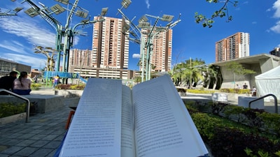 A book is open in the foreground, held against a background of an urban park. Tall buildings and unique metal structures with solar panels are visible. The park has greenery, benches, and people sitting and walking around under a clear blue sky.