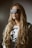 A person with long, wavy blonde hair is wearing oversized, reflective sunglasses with a grid pattern. The person is dressed in a white top featuring bold, curved black lettering and a black belt with multiple buckles. The background is a muted, dark gradient, adding to the overall dramatic and stylish appearance.