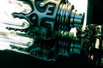 A close-up view of a decorative metal spray nozzle with intricate, ornamental patterns. The nozzle is attached to a chain and has reflective surfaces that catch the light, creating a dramatic contrast. The background is dark, adding to the mysterious and artistic feel of the image.