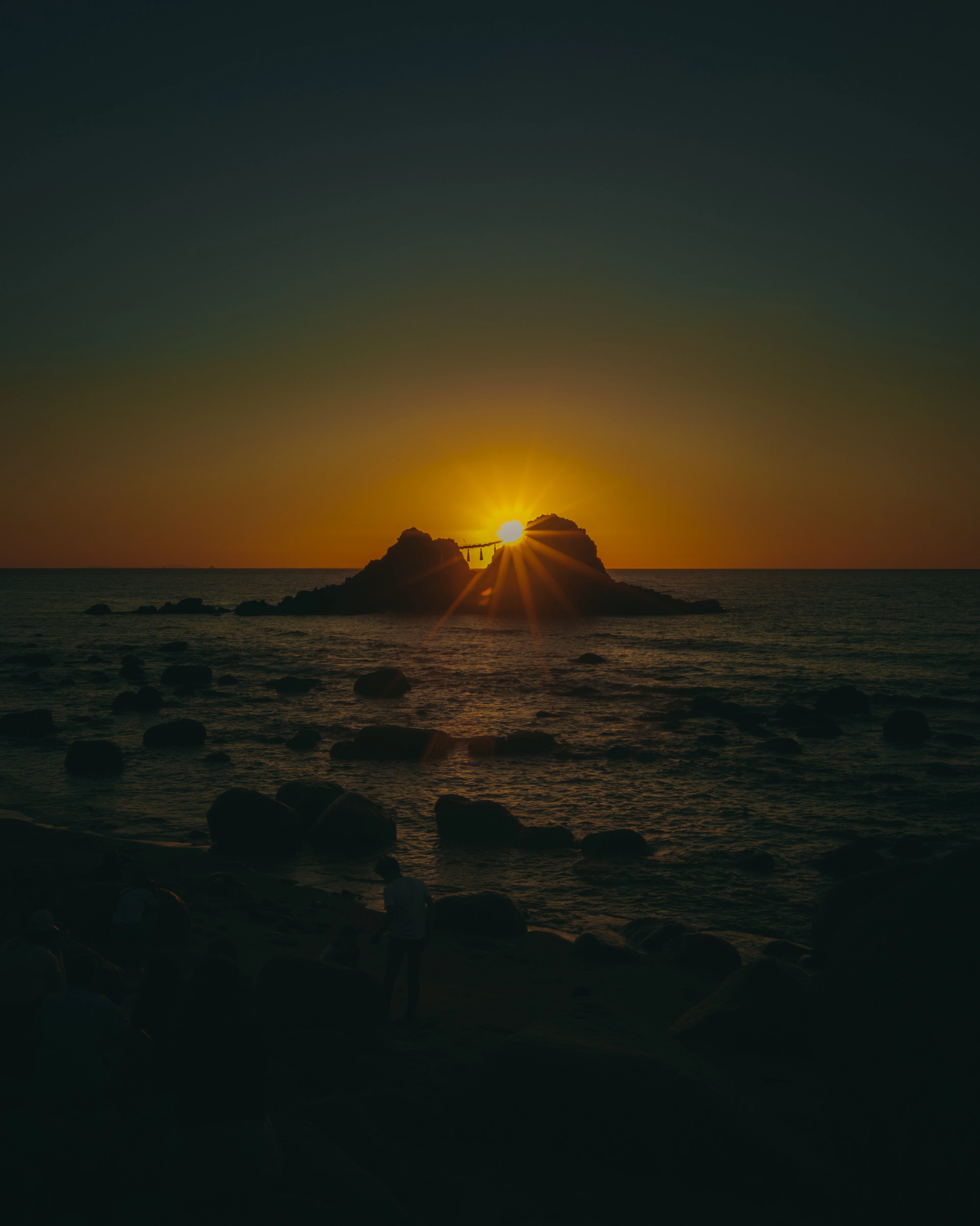 Silhouetted rocks holding a torii gate glow against an orange ocean sunset as a figure watches from the shore.