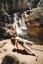 A person is practicing a yoga pose on a rock platform next to a small waterfall surrounded by lush greenery. The scene is sunlit, creating contrasting shadows and highlights on the person's clothing and the natural surroundings.