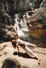 A person is practicing a yoga pose on a rock platform next to a small waterfall surrounded by lush greenery. The scene is sunlit, creating contrasting shadows and highlights on the person's clothing and the natural surroundings.