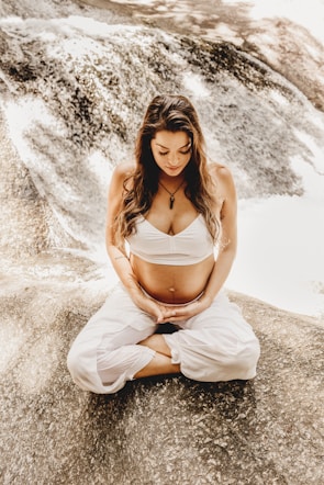 A woman in a meditative posture, sitting cross-legged on a rock beside a flowing stream. She is wearing a white top and pants, with her hands gently resting on her belly.