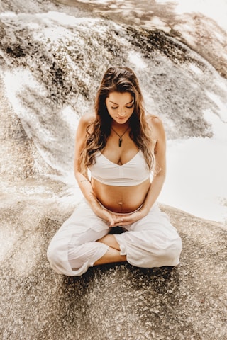 A woman in a meditative posture, sitting cross-legged on a rock beside a flowing stream. She is wearing a white top and pants, with her hands gently resting on her belly.