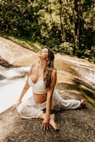 A woman is practicing yoga on a rock beside a serene stream in a lush forest setting. She is wearing a white top and pants, looking relaxed and at ease.