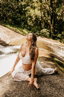 A woman is practicing yoga on a rock beside a serene stream in a lush forest setting. She is wearing a white top and pants, looking relaxed and at ease.