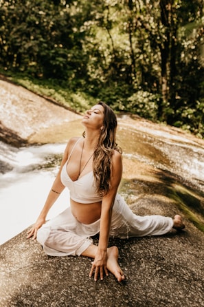 A woman is practicing yoga on a rock beside a serene stream in a lush forest setting. She is wearing a white top and pants, looking relaxed and at ease.