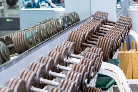 A row of metal dumbbells neatly arranged on a rack in front of a mirrored wall. The background shows gym equipment and reflections. The setting is clean and organized, emphasizing functional fitness spaces.