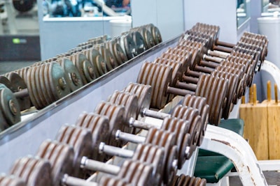 A row of metal dumbbells neatly arranged on a rack in front of a mirrored wall. The background shows gym equipment and reflections. The setting is clean and organized, emphasizing functional fitness spaces.