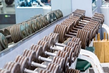 A row of metal dumbbells neatly arranged on a rack in front of a mirrored wall. The background shows gym equipment and reflections. The setting is clean and organized, emphasizing functional fitness spaces.