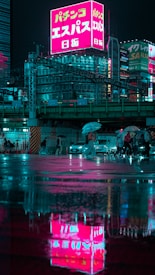 A vibrant urban scene at night featuring illuminated buildings with neon signs in Japanese, reflecting in rain-soaked streets. People with umbrellas cross the road under the glow of the lights. The atmosphere is moody yet vivid with reflections enhancing the visual depth.