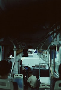The inside of a bus is visible, with several empty seats and a view out the front window. A person is seated in the driver's seat, and the road ahead is visible through the windshield. The interior lighting is dim, and there are yellow handles hanging from the ceiling for standing passengers.