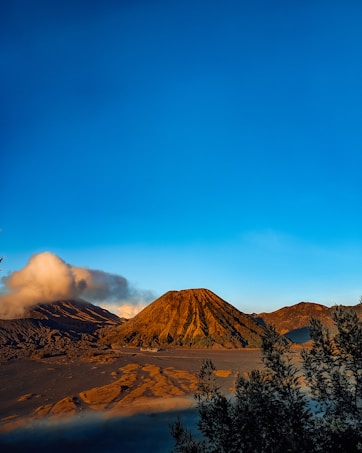 A landscape featuring a prominent volcano with a rugged, brown surface under a bright blue sky. Adjacent to the volcano, another peak emits a plume of white smoke. The foreground is framed by dark green foliage, adding depth to the composition.